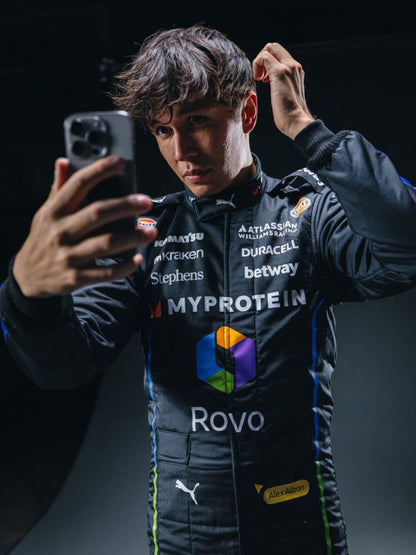 Alexander Albon, wearing a black 2025 Williams Las Vegas GP F1 racing suit with various sponsor logos, is taking a selfie - GPX WEAR