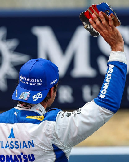 Carlos Sainz wearing a white 2025 Williams United States GP F1 racing suit and cap with 'Arrows' branding, holding a trophy against a blurred background - GPX WEAR