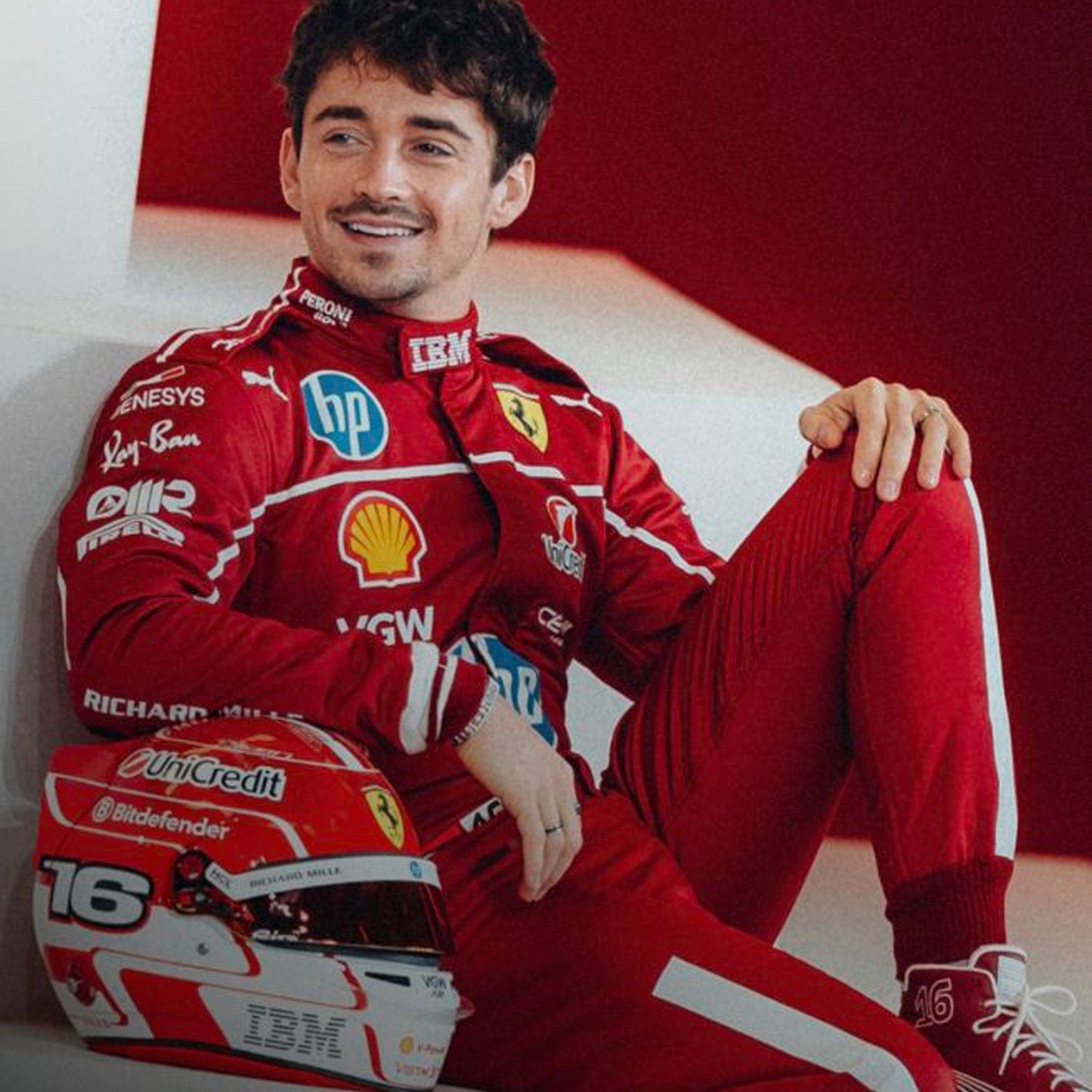 Charles Leclerc is wearing a red 2025 Ferrari F1 racing suit with various sponsor logos, sitting next to a helmet - GPX WEAR