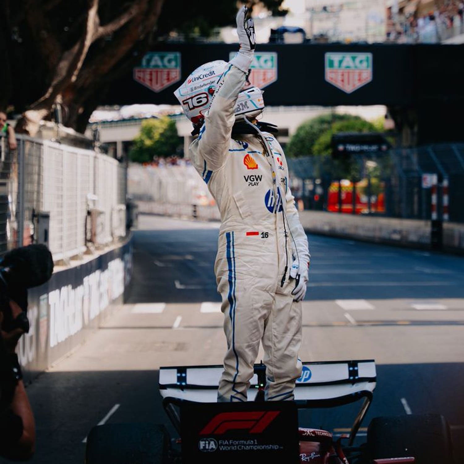 2025 Charles Leclerc Monaco GP f1 Racing Suit with sponsor logos, standing on a podium with a Formula 1 car in the background.