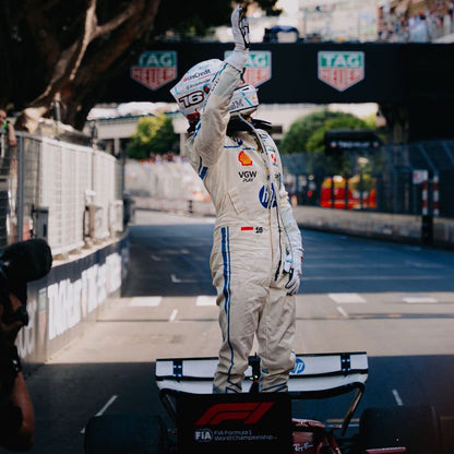 2025 Charles Leclerc Monaco GP f1 Racing Suit with sponsor logos, standing on a podium with a Formula 1 car in the background.