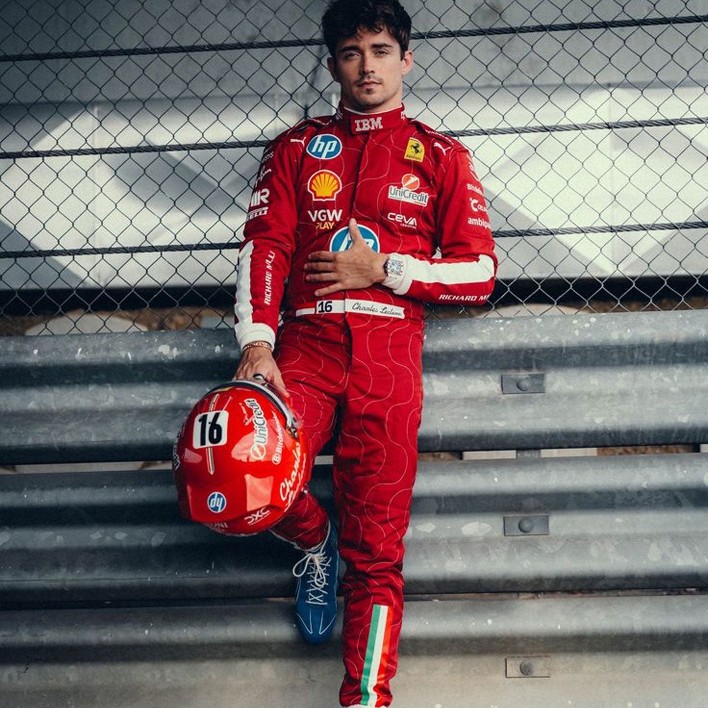 Charles Leclerc wearing a red 2025 Ferrari Monza GP F1 racing suit holding a helmet with a fence - GPX WEAR