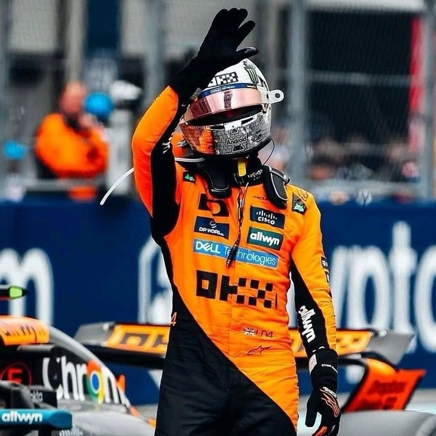 Lando Norris wearing an orange and black McLaren 2025 F1 racing suit and helmet waving at a race track with a blurred background - GPX WEAR
