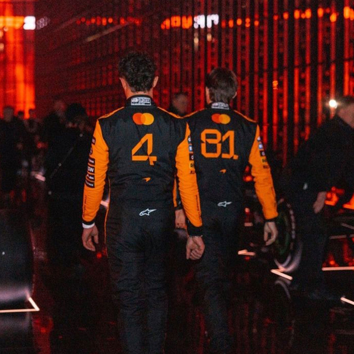 Back view of two individuals, Lando Norris and Oscar Piastri, in 2025 McLaren F1 racing suits with visible numbers walking in a dark setting - GPX WEAR